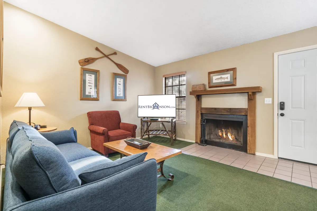 A warmly lit living room centered around a glowing stone fireplace, with a plush sofa and armchair inviting relaxation. A television is set nearby, and decorative paddles on the wall add a nostalgic lakeside touch. Large windows let in natural light and offer a glimpse of wooded views.