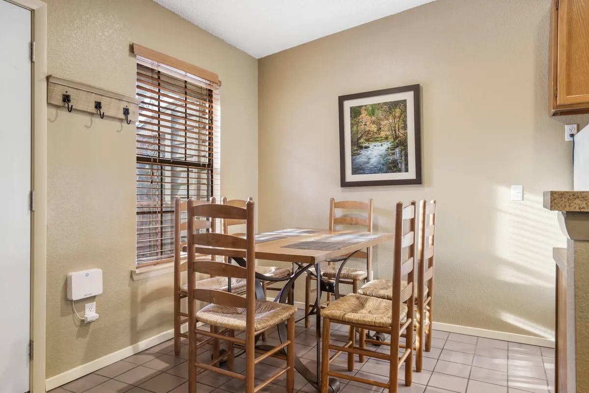 A comfortable dining area with a large table, chairs, wall art, and a window letting in natural light. The scene conveys a sense of gathering and lingering conversation.