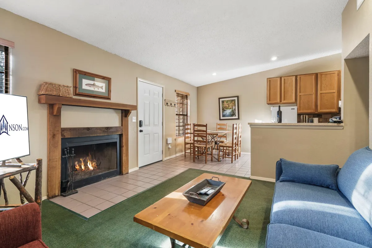 A warm, inviting interior featuring a stone fireplace, a cozy sofa, a dining table ready for board games or shared meals, and an open kitchen area. The scene gives off classic camp lodge vibes, perfect for gatherings, story-sharing, or cozy evenings after outdoor adventures.
