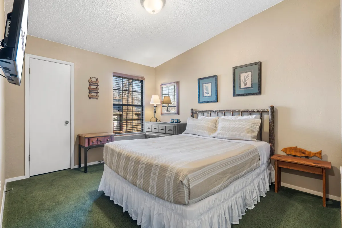 A warmly lit bedroom featuring a queen bed with plush bedding, a nightstand with a lamp, a window that lets in natural sunlight, and tasteful artwork above the bed. The view through the window hints at wooded surroundings, enhancing the tranquil, camp-inspired vibe.