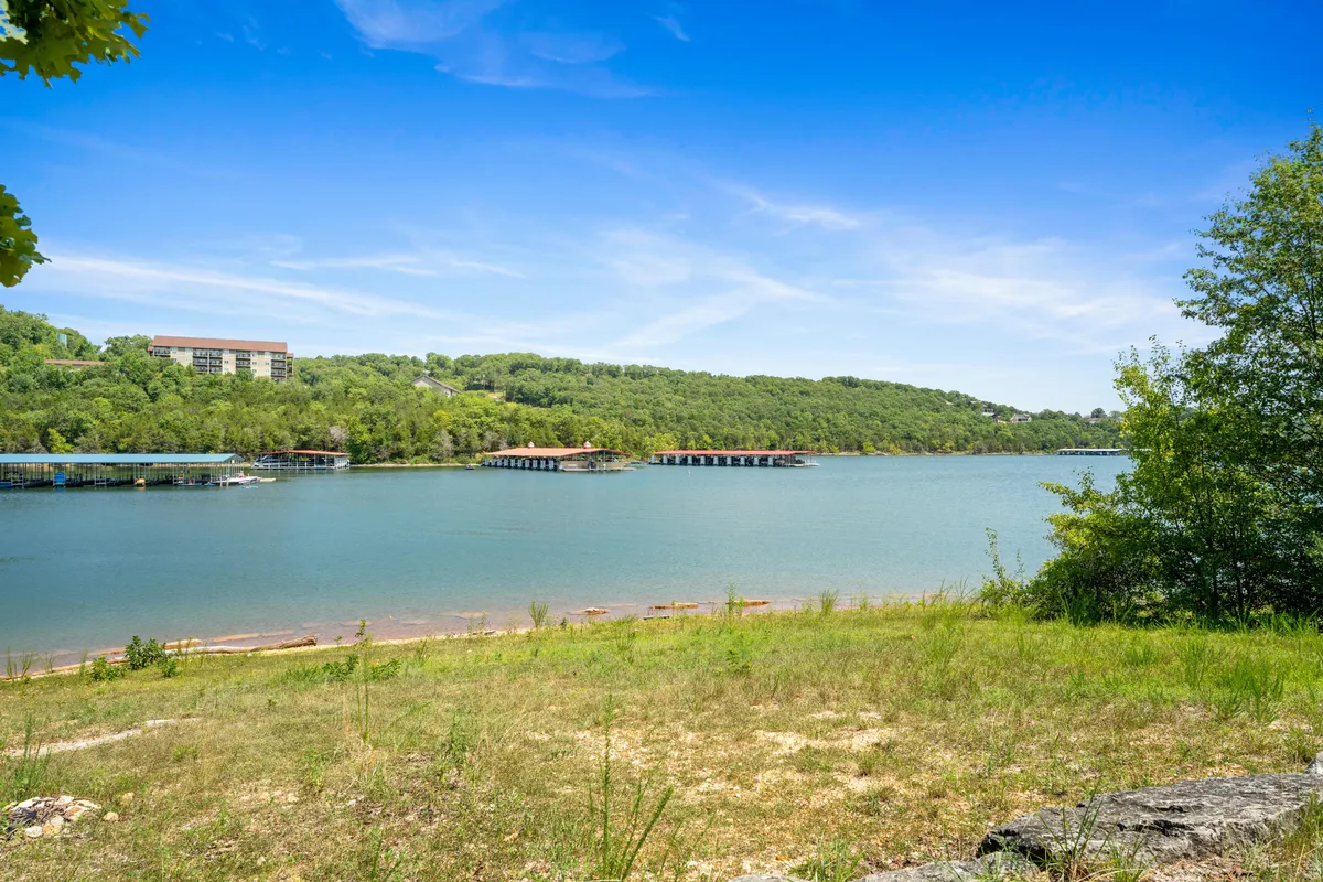 A panoramic view of calm lake waters surrounded by lush green hills and dotted with boathouses under a clear sky. The scene emanates tranquility and adventure, suggesting opportunities for lakeside picnics, scenic walks, and water-based activities.
