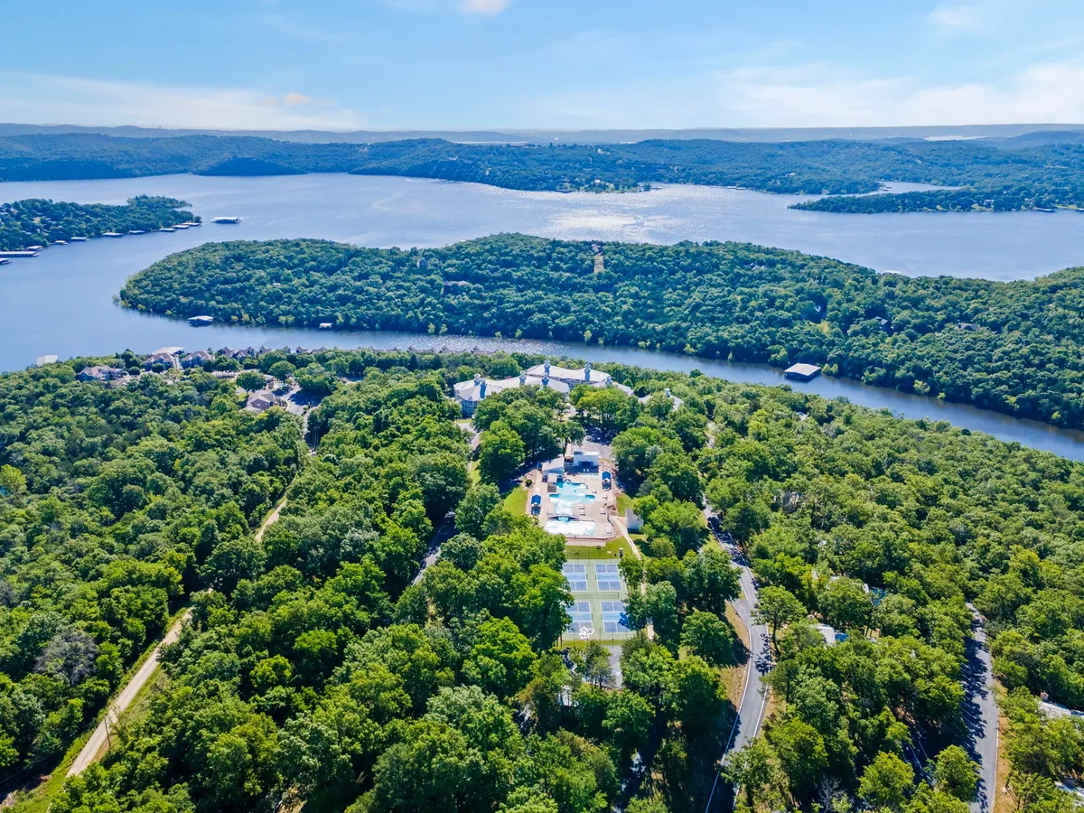 An exterior view showcasing an expansive water body bordered by a lush green wooded area, with residential structures and recreational facilities in clear view. The scene suggests a tranquil and inviting atmosphere for outdoor enjoyment.