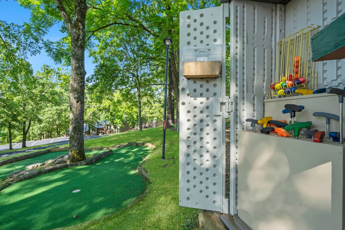 A playful mini-golf putting area accented by green grass, colorful tools, and surrounding trees. The scene evokes family fun, friendly competition, and outdoor leisure in a natural setting.