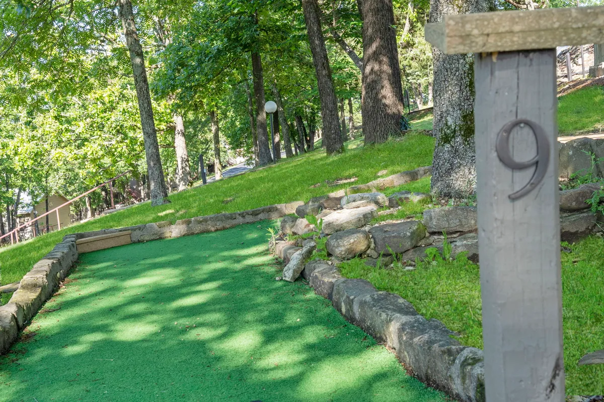 This exterior image captures a vibrant putting green nestled among mature trees and rock borders. The cheerful atmosphere is highlighted by a numbered post, inviting families to engage in friendly mini-golf competitions surrounded by nature.