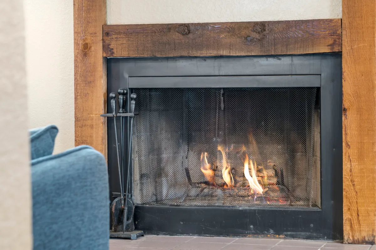 A close-up of a rustic fireplace with a wooden mantel and fire tools arranged nearby. The fire glows warmly, creating a focal point for gatherings, reading, or unwinding after outdoor adventures.