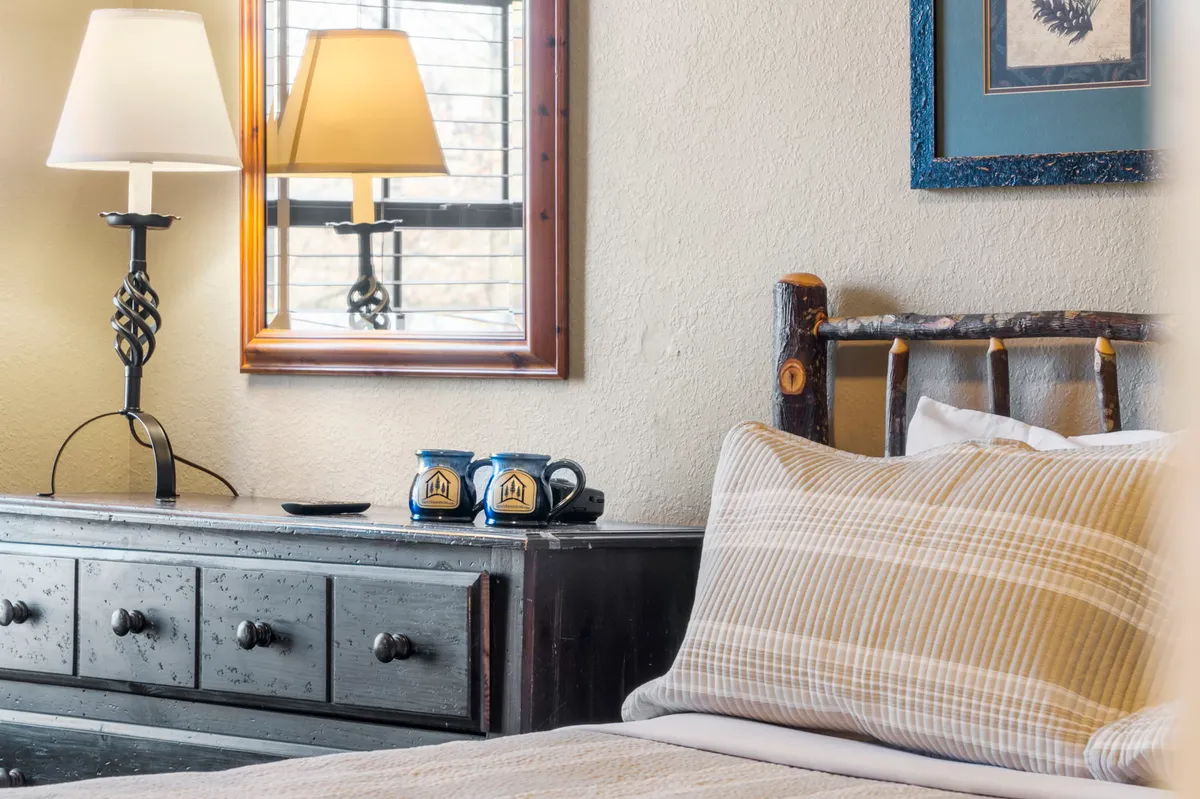 A comfortable bedroom with a wooden dresser, a queen bed with soft linens, a cozy lamp for evening reading, and coffee mugs placed on the dresser—suggesting a relaxing morning routine. Natural light filters in, creating a warm, inviting environment.