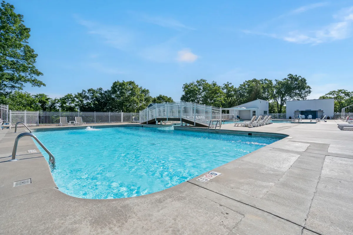 A beautifully landscaped outdoor swimming pool area featuring loungers, a charming bridge, and abundant greenery. The scene is lively yet leisurely, with families and guests enjoying the water and poolside amenities, highlighted by the inviting blue pool under the open sky.